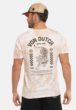 Homme portant un t-shirt tie-dye beige avec un motif tigre rugissant et le texte "Von Dutch" au dos, casquette de baseball beige et noire, short noir.