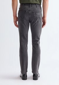 Moški v sivoh tesno prilegajočih kavbojkah Wrangler in črnih elegantnih čevljih, prikazan od zadaj na preprostem svetlem ozadju.