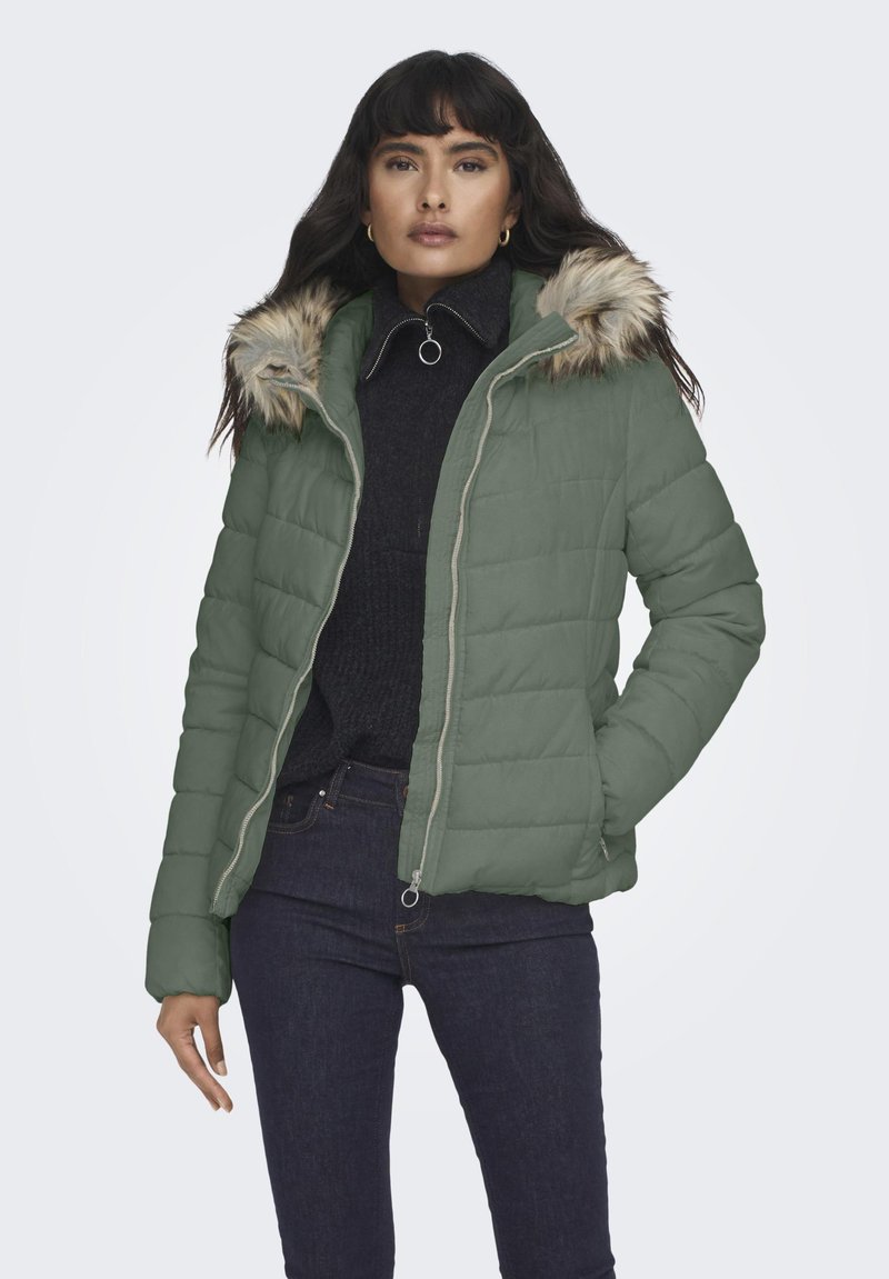 Grønn quiltet pufferjakke med faux fur hette, glidelås og lange ermer. Båret over en svart genser og paret med mørke jeans.