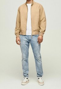 Chaqueta bomber de cremallera color marrón claro con puños y dobladillo acanalados, combinada con jeans de color azul claro y zapatillas beige con acentos blancos.