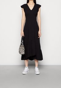 Nainen mustassa midi-mekoissa, jossa on röyhelöhihoja, pitää zebratulostettua käsilaukkua ja käyttää valkoisia platform-tennareita, seisten harmaalla lattialla.