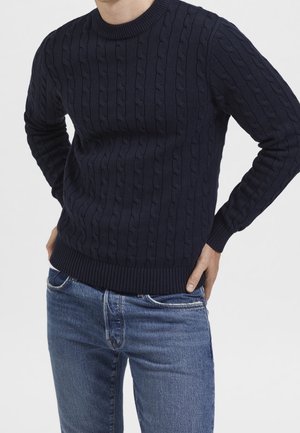 Man wearing navy blue cable-knit sweater and blue jeans, hands resting on hips against a plain light background.