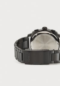 Montre en acier inoxydable noir avec un bracelet texturé, des maillons polis et des accents mats. Elle présente un boîtier rond et le logo de la marque gravé sur le fermoir.