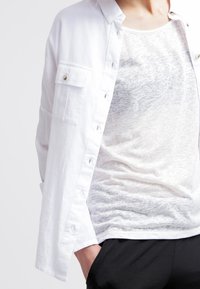 Vit skjorta med knäppning och en bröstficka, kombinerad med en genomskinlig vit tanktopp med subtil textur och lagerutseende, bärs med svarta byxor.