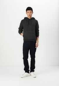 Sudadera de forro polar negra con un bolsillo canguro, combinada con jeans de mezclilla oscuros y zapatillas blancas. El modelo está de pie mirando hacia adelante frente a un fondo blanco.