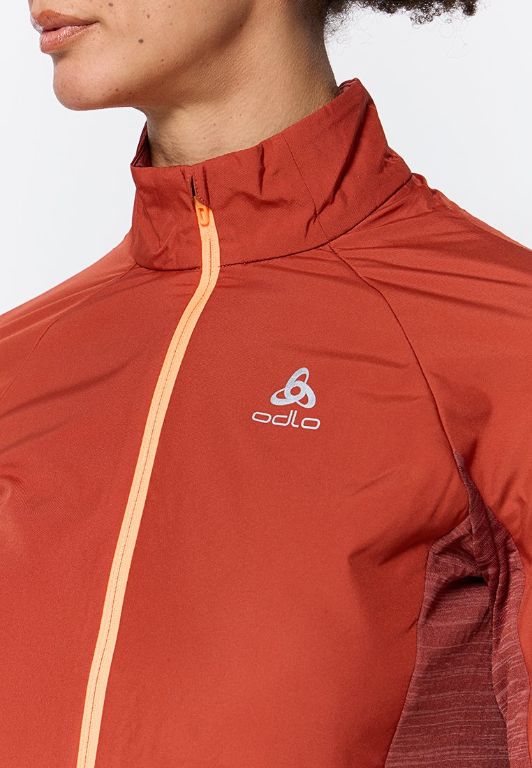 Rote Sportjacke mit orangefarbenem Reißverschluss, hohem Kragen und Odlo-Logo. Sie verfügt über eine glatte Textiloberfläche mit kontrastierenden strukturierten Einsätzen.