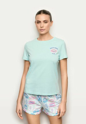 Kobieta ubrana w miętowy t-shirt "Salty Soul Club" i białe szorty z kolorowymi ilustracjami puszek sardynek, stojąca na jednolitym tle.