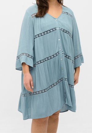 Femme portant une robe ample bleu clair avec des bandes de dentelle diagonales et des boutons sur le devant, debout les bras détendus le long du corps.