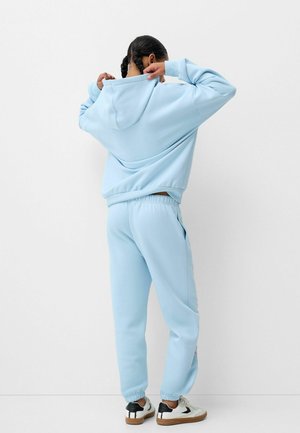 Persoon met gevlochten haar draagt een lichtblauwe hoodie en bijpassende joggingbroek, doet de capuchon recht, staat voor een witte muur, witte sneakers met zwarte details.