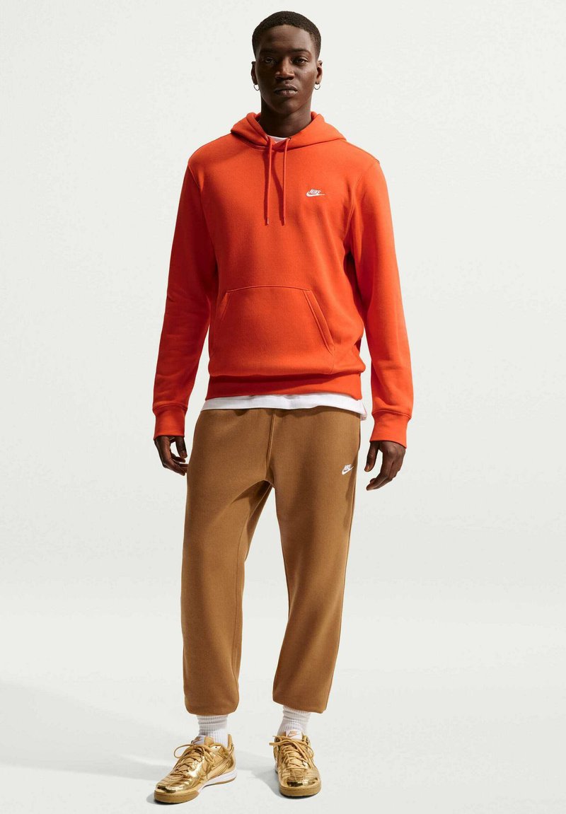 Sudadera con capucha de forro polar roja con bolsillo tipo canguro y logo blanco, combinada con pantalones jogger marrones. Zapatillas doradas con cordones y calcetines blancos hasta la pantorrilla.