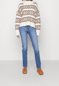 Suéter blanco tejido con patrones geométricos marrones, combinado con jeans azules y zuecos color beige. El suéter presenta un cuello y puños acanalados.