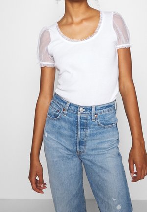 Mujer que lleva una camiseta blanca con mangas globo transparentes y vaqueros azules de talle alto con un ligero desgaste.