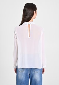 Blusa bianca semi-trasparente con maniche lunghe, polsini con bottoni, un colletto piccolo e un'apertura a forma di chiave sul retro. Abbinata a jeans blu.