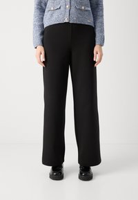 Pantalon large noir en matériau lisse avec une finition soignée, assorti à des chaussures à plateforme noires, porté avec un cardigan texturé bleu.