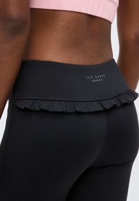 Κοντινό πλάνο ενός ατόμου που φοράει μαύρο κολάν με ψηλή μέση, διακοσμημένη με βολάν και λογότυπο «Ted Baker Sport» στη μέση.