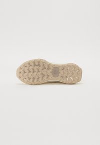 Lys beige såles med teksturert gummigrep, med et sikksakk-mønster og et sentralt emblem. Flat, robust design for godt grep.