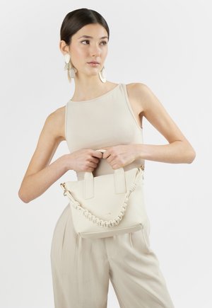 Giovane donna con top beige senza maniche e pantaloni, che tiene una borsa crema con manico intrecciato, indossa grandi orecchini rotondi, guarda di lato.