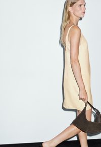 Donna in un vestito beige senza maniche che cammina, tenendo una borsa in camoscio marrone scuro, su uno sfondo bianco semplice.