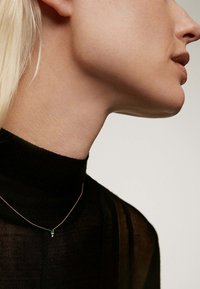 Collar de oro con un pequeño colgante de gema verde. El modelo lleva un suéter negro de cuello alto, mostrando el collar en el cuello.
