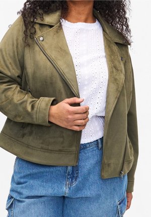 Personne aux cheveux bouclés portant une veste en daim vert olive, un chemisier blanc ajouré et un jean denim taille haute bleu, ajustant la fermeture éclair de la veste.
