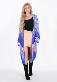 Long cardigan en tricot bleu et blanc avec un motif géométrique, porté sur un haut noir et une jupe beige, associé à des bottes noires montantes.