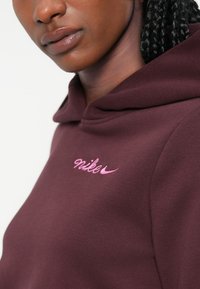 Bordo kapuca s izvezenim ružičastim "Nike" logotipom. Ima kapu s vezicom i mekanu, glatku teksturu. Pogled izbliza na vratni izrez i kapucu.