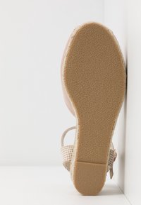 Sandale avec semelle en caoutchouc texturée, talon compensé enveloppé de jute et bride tissée dans des tons beige clair, placée debout contre un coin blanc.