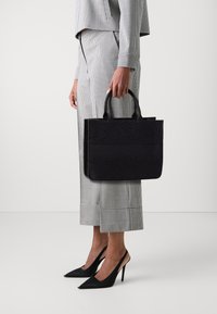 Borsa a spalla nera con motivo, dotata di doppi manici; forma strutturata con texture diverse, tenuta da una persona in pantaloni a quadri e scarpe con tacco.