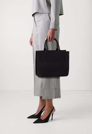 Borsa a spalla nera con motivo, dotata di doppi manici; forma strutturata con texture diverse, tenuta da una persona in pantaloni a quadri e scarpe con tacco.