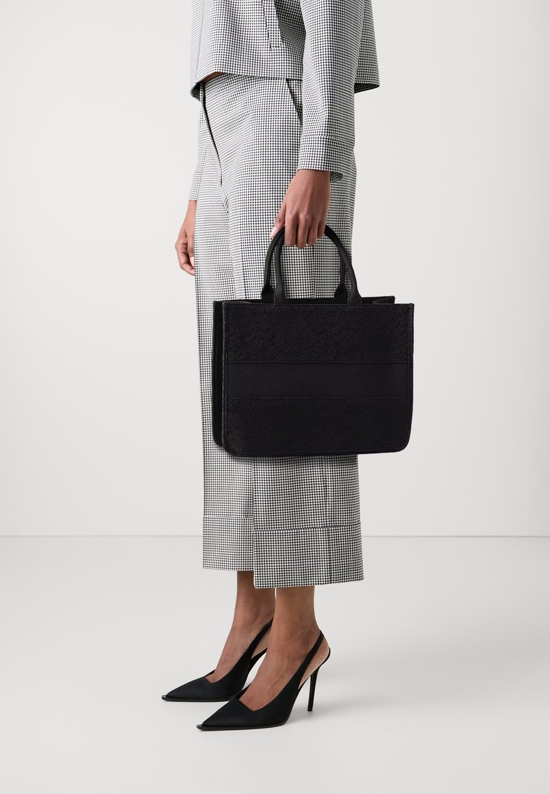 Borsa a spalla nera con motivo, dotata di doppi manici; forma strutturata con texture diverse, tenuta da una persona in pantaloni a quadri e scarpe con tacco.