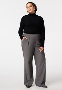 Vrouw in een zwarte coltrui, grijze wijde broek, gouden ringsieraden en zwarte puntige schoenen met gouden studs, staand met één hand in haar zak.