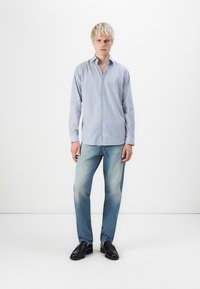 Blau-weiß gestreiftes Button-Up-Hemd aus leichtem Stoff, kombiniert mit hellblauen Jeans und schwarzen Slippern. Das Model steht vor einem weißen Hintergrund.
