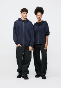 Navy blue zip-up hoodies made of soft fabric, paired with baggy black denim pants. Both individuals wear black footwear, with relaxed fits.