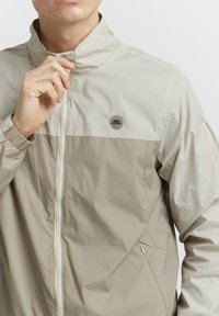 Lätt, beige vindjacka med hög krage, färgblockerad design, dragkedjestängning och en logotyp på bröstet. Slät struktur.