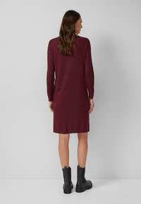 Frau mit langen braunen Haaren, die ein knielanges burgunderfarbenes Strickkleid und schwarze Lederstiefeletten trägt und vor einer schlichten grauen Wand steht.