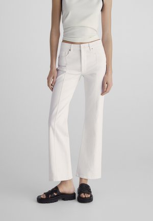 Jean flare blanc avec taille haute, poches avant et coutures contrastées. Associé à des sandales compensées noires en toile. Texture de tissu lisse.