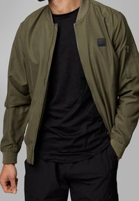 Homme portant une veste bomber vert olive ouverte sur un t-shirt noir et un pantalon noir sur fond gris.
