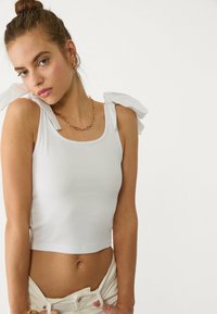 Bijeli rebrasti crop top s širokim tkanim mašnama na ramenima i okruglim dekolteom, u kombinaciji s svijetlim donjim dijelom.