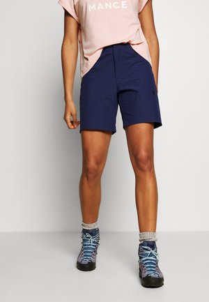 Person in marineblauen Shorts, pinkem Hemd mit hochgekrempelten Ärmeln, grauen Socken und violetten Wanderschuhen, stehend vor einem schlichten hellen Hintergrund.