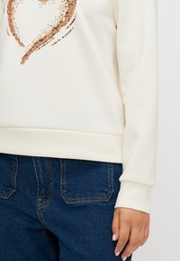 Kermakiven huppari, jossa on sydänkuvio rose gold -paljeteista. Varustettuna ribbitetyillä hihoilla ja rentoa istuvuutta. Yhdistetty tummansinisiin farkkuihin.