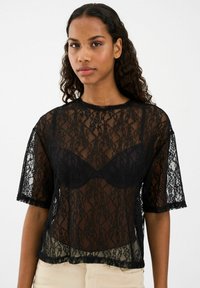 Melnas mežģīņu blūze ar īsām piedurknēm, caurspīdīgu dizainu, ziedu rakstiem un viļņotu apakšmalu, kārtota virs melna braleta.