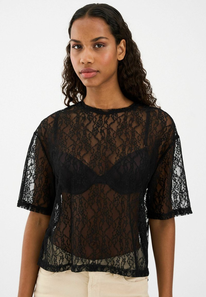 Melnas mežģīņu blūze ar īsām piedurknēm, caurspīdīgu dizainu, ziedu rakstiem un viļņotu apakšmalu, kārtota virs melna braleta.