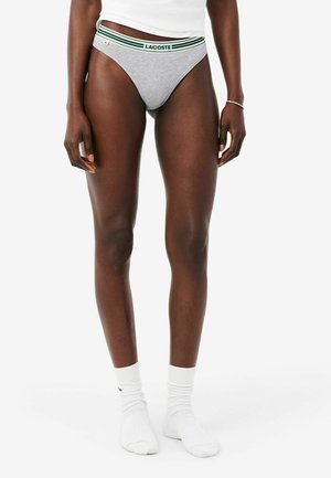 Frau trägt graue Lacoste-Slips mit grün gestreiftem Bund, weiße Socken und weißes Top, stehend vor weißem Hintergrund.