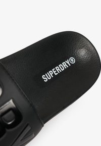 Sandal nera a infradito realizzata in materiale sintetico testurizzato. Presenta un logo "SUPERDRY®" bianco in evidenza sulla fascia. Design con suola piatta.