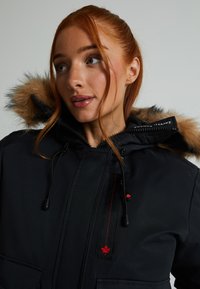 Fekete parkau fur bélelő kapucnival, cipzárral, húzózsinórokkal és piros juharlevél díszítéssel. Puha tapintású, elülső zsebekkel rendelkezik.