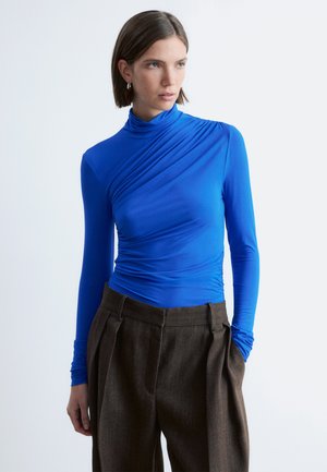 Vrouw met kort bruin haar, gekleed in een felblauwe top met lange mouwen en draperdetails, en hoge, losse bruine broek tegen een witte achtergrond.