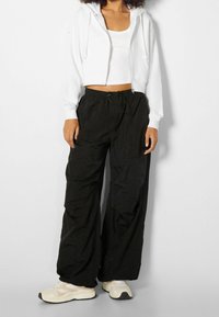 Donna che indossa una felpa bianca a zip corta, un top bianco, pantaloni neri larghi con lacci e sneakers beige, in piedi contro uno sfondo neutro.