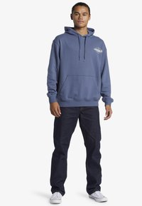 Modrá mikina s kapucňou, predným vreckom, šnúrkami a detailom loga; skombinovaná s tmavými džínsami a bielymi teniskami.