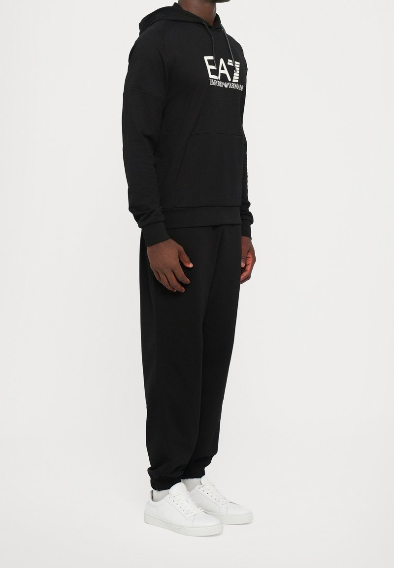 Homme portant un sweat à capuche noir EA7 Emporio Armani, un pantalon noir et des baskets blanches, debout sur un fond blanc.