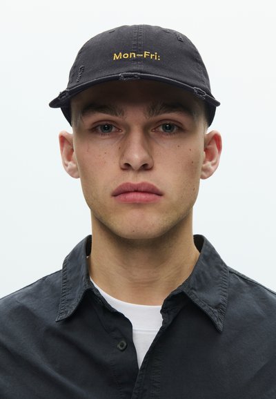 Μαύρο καπέλο με φθαρμένες λεπτομέρειες και κίτρινη κεντημένη επιγραφή «Δευ-Παρ» στο μπροστινό μέρος, φορεμένο με μαύρο πουκάμισο με κουμπιά πάνω από λευκό μπλουζάκι.